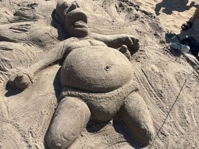 A sand sculpture of a large reclining figure on a beach, with beachgoers standing around it.