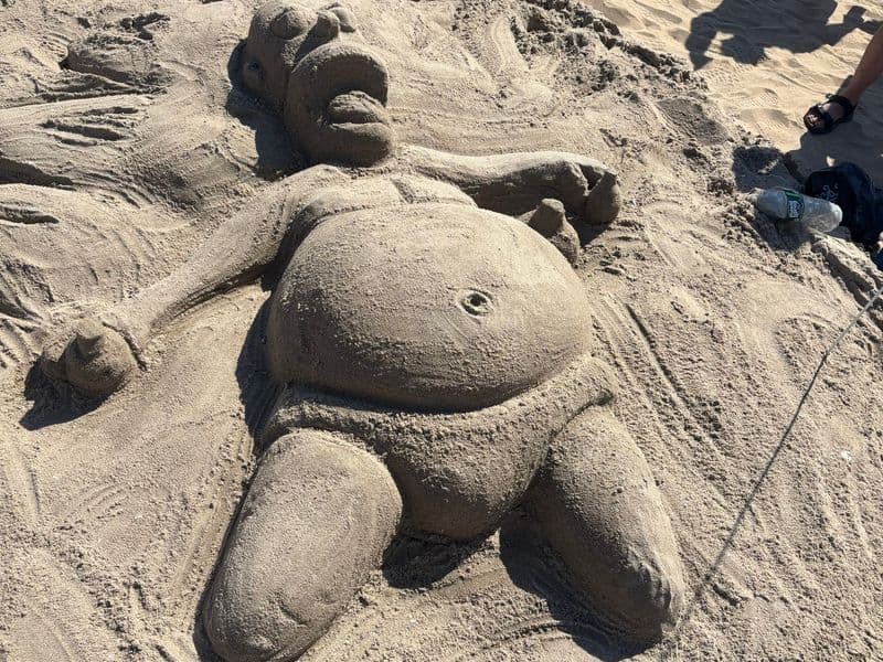 A sand sculpture of a large reclining figure on a beach, with beachgoers standing around it.